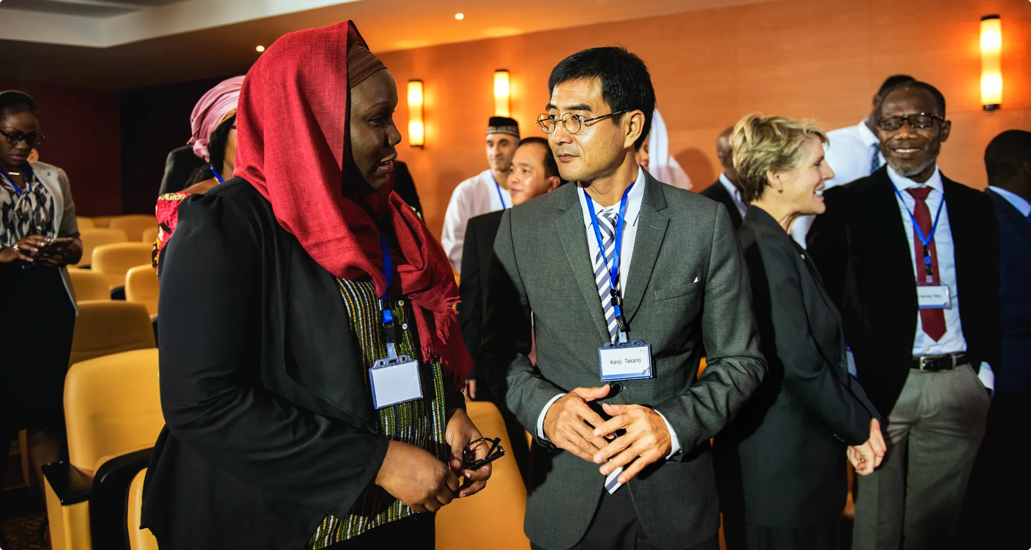 Two delegates with lanyards talk in the auditorium before a session